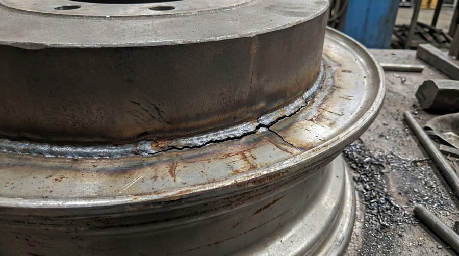 An animation-style image showing arrows of force flowing into a wheel disc, being deflected by the thick disc, and concentrating intensely at the weld line.